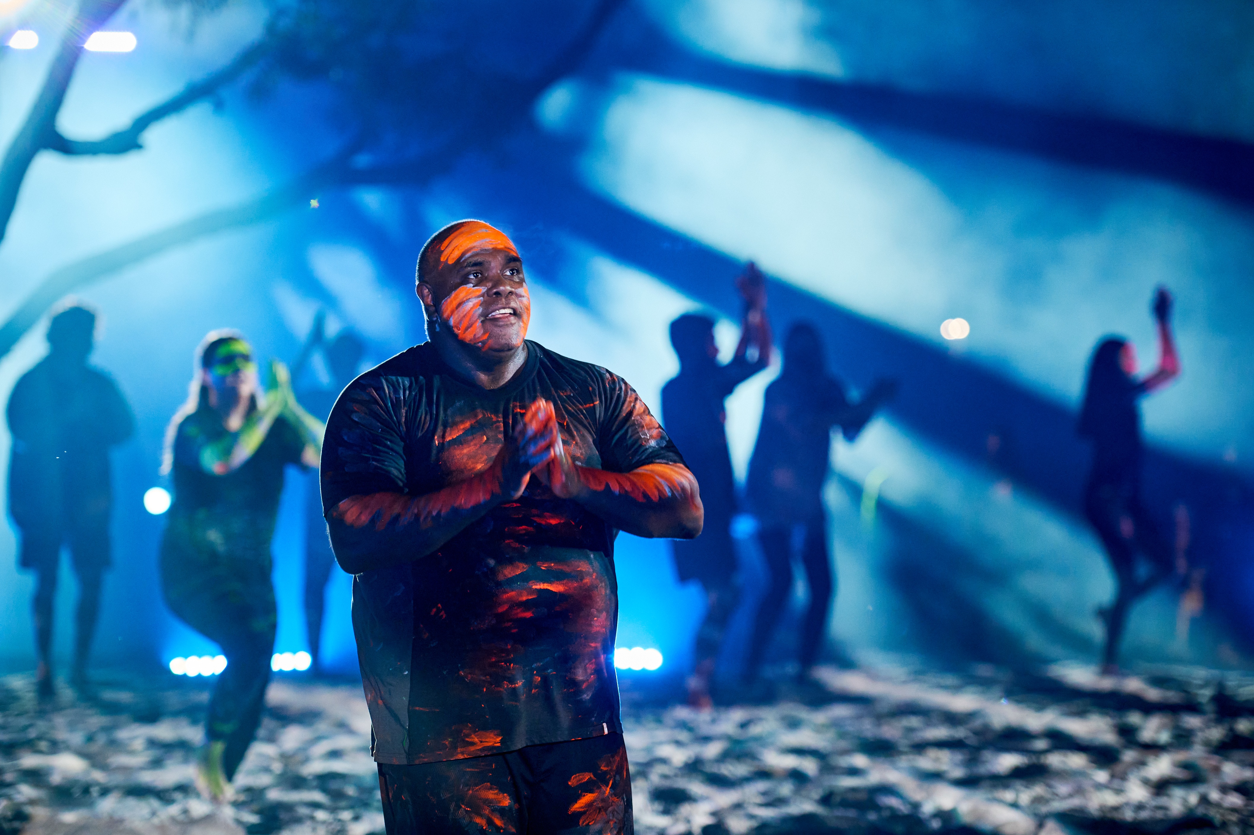 A man in orange and blue body paint on stage with people silhouetted in background