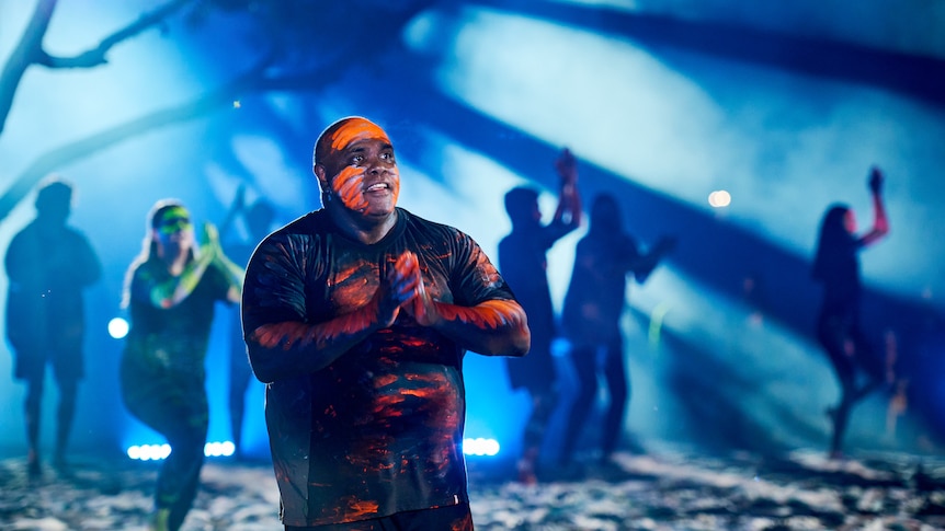 A man in orange and blue body paint on stage with people silhouetted in background