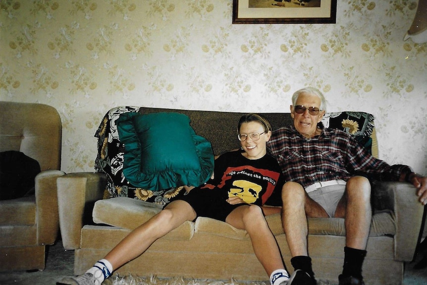 A young boy and his grandfather sit on a lounge in a 1980s unit. Floral wallpaper is in the background.