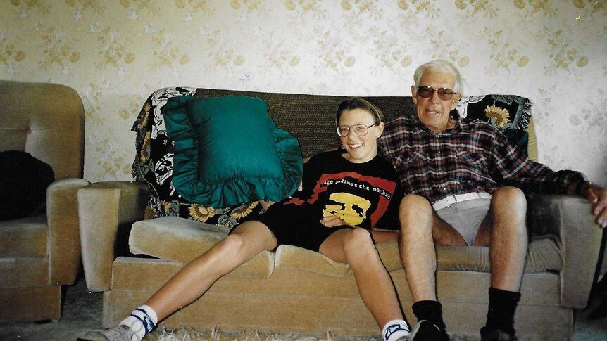 A young boy and his grandfather sit on a lounge in a 1980s unit. Floral wallpaper is in the background.