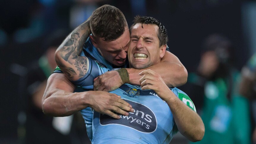 Mitchell Pearce is hugged from behind by a teammate and he reaches up and hugs back with a smile on his face.