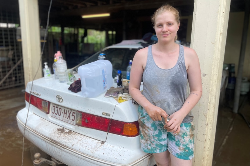 A woman in a grey singlet leans against her car with rubbish and mud behind her