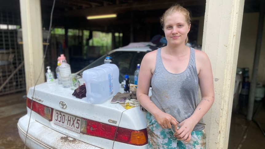 A woman in a grey singlet leans against her car with rubbish and mud behind her.
