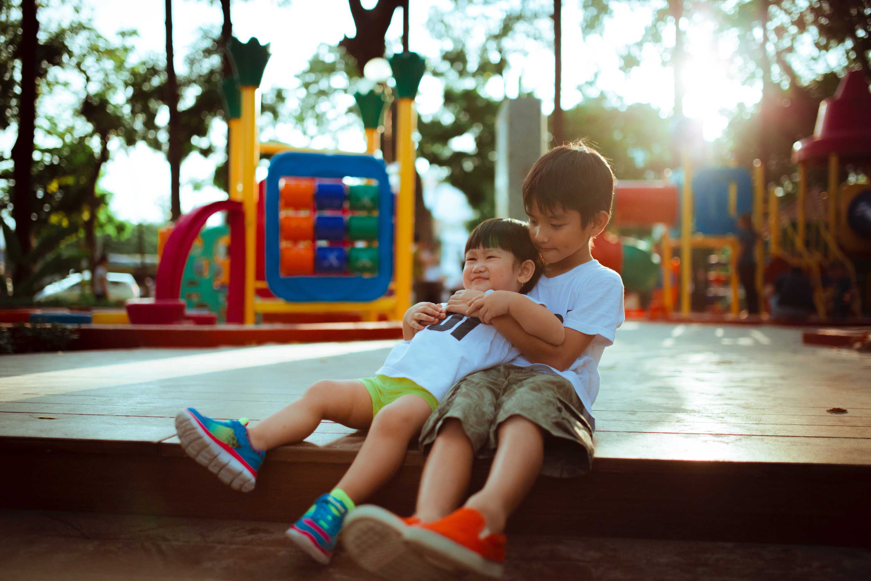 Two young boys sitting at a playground, the older one is holding the younger one