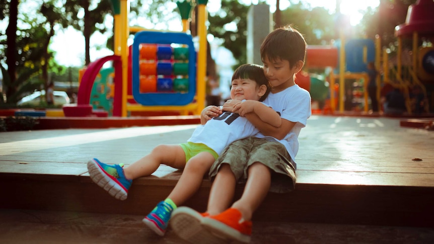 Two young boys sitting at a playground, the older one is holding the younger one