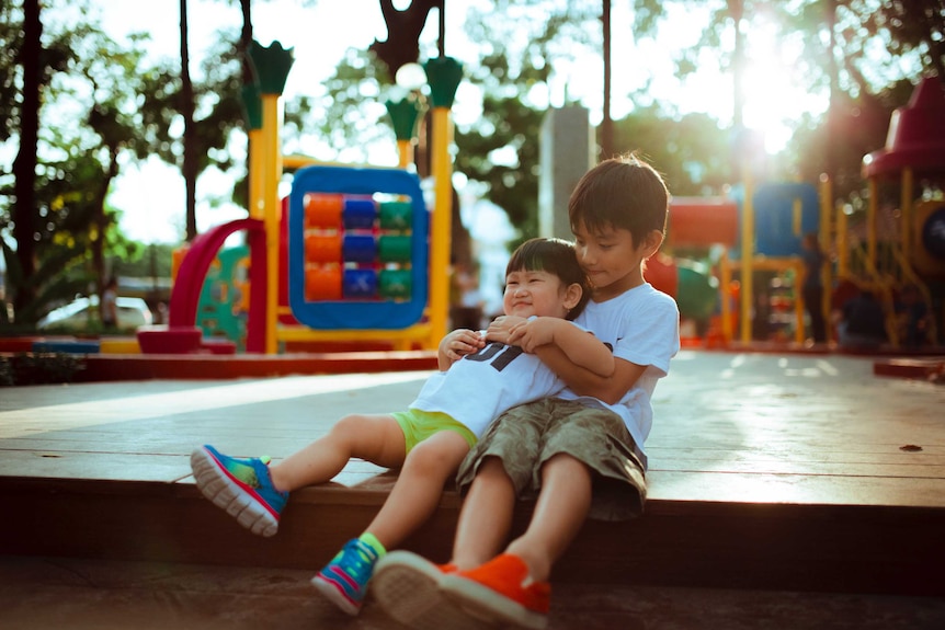Two young boys sitting at a playground, the older one is holding the younger one