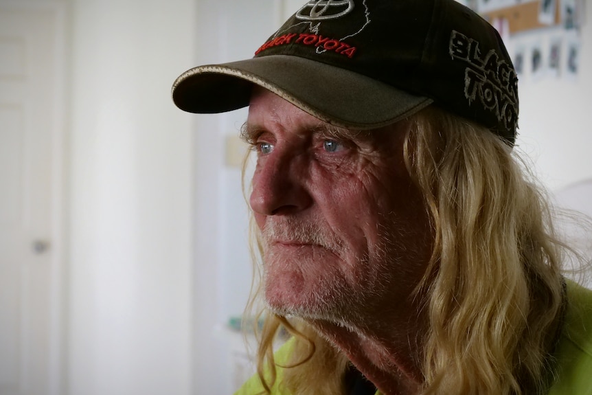 A close up of a man with long blonde hair wearing a cap, he has a sad expression