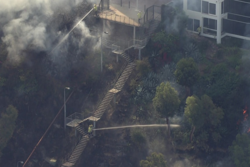Firefighters stand on an outdoor staircase as they spray water into bushland on fire 
