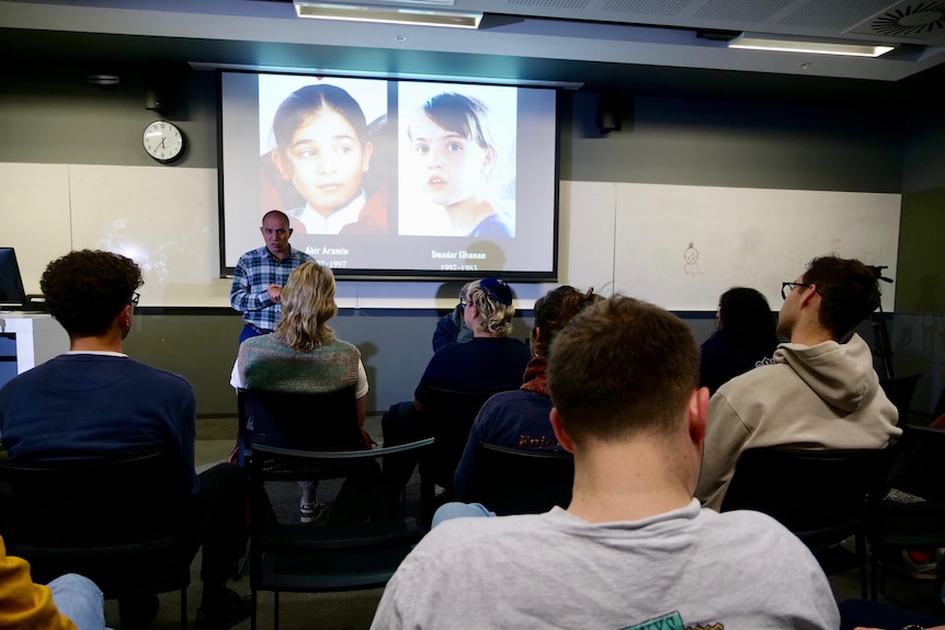 A man stands in front of a room of people - the faces of two young girls are show on a projector screen