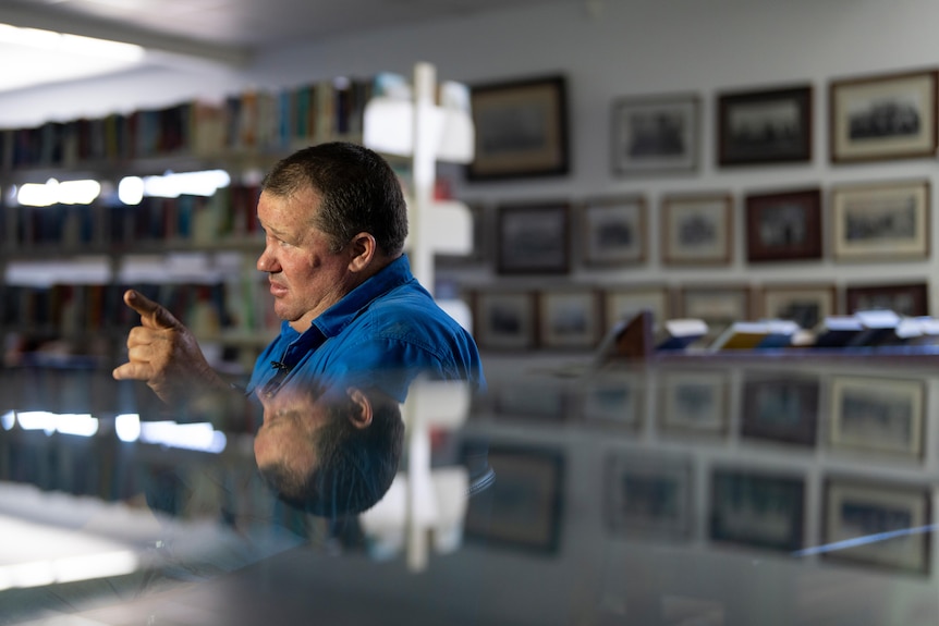 A man in a blue shirt sitting behind a reflective desk. He's pointing his finger