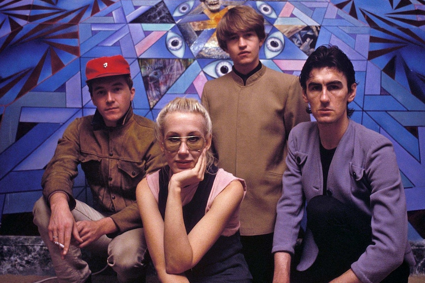 A band of four young people in the 1980s sit together behind a purple backdrop.