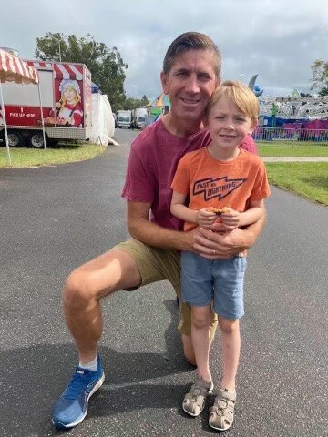 Man with neat hair crouches down a young boy and embraces him. Carnival rides are seen in the background