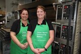Two women in green aprons stand in front of an oven in a bakery.