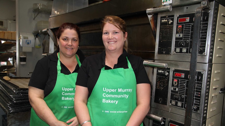 Two women in green aprons stand in front of an oven in a bakery.