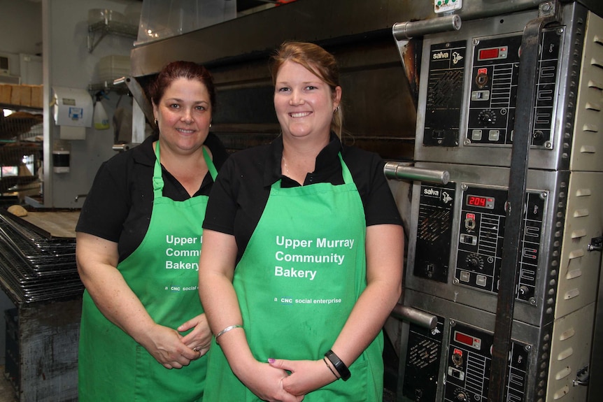 Two women in green aprons stand in front of an oven in a bakery.