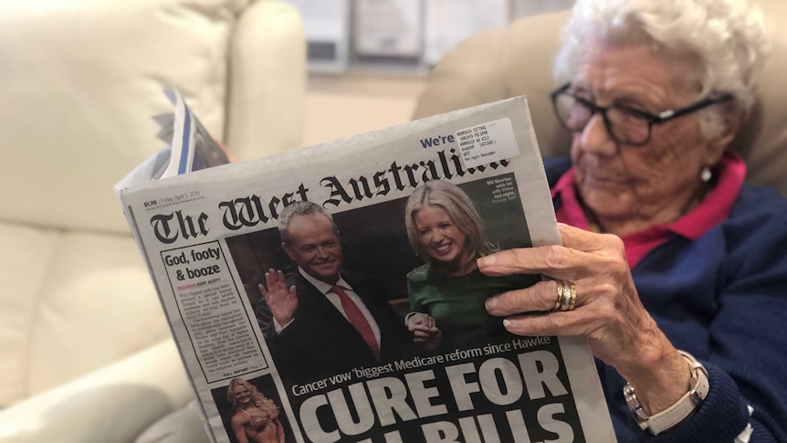 An elderly woman sits in an armchair at a care home reading a newspaper