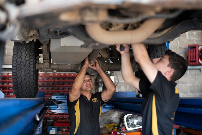 Two men working on the underside of a car which is on a hoist above their heads.