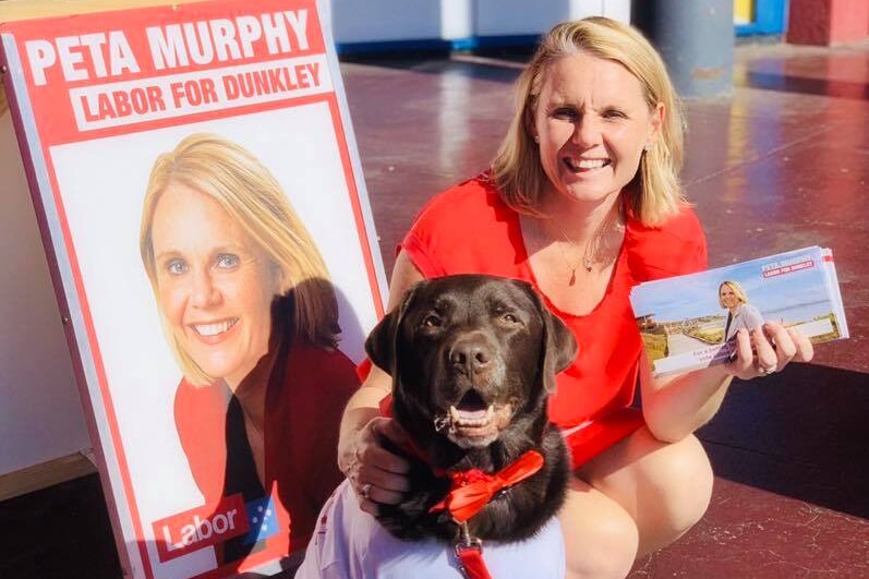 A woman smiles as she sits next to a dog in front of her campaign poster holding leaflets.