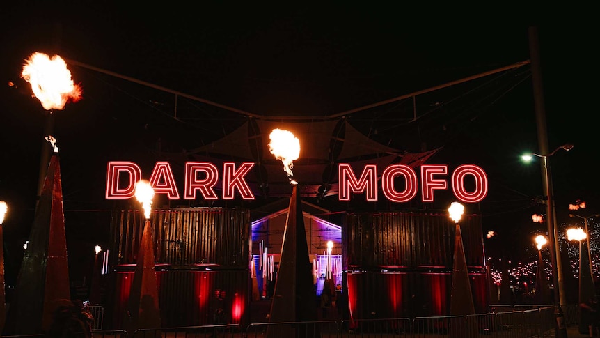 Neon red lights reading "Dark Mofo" amid pyramids shooting out fire.
