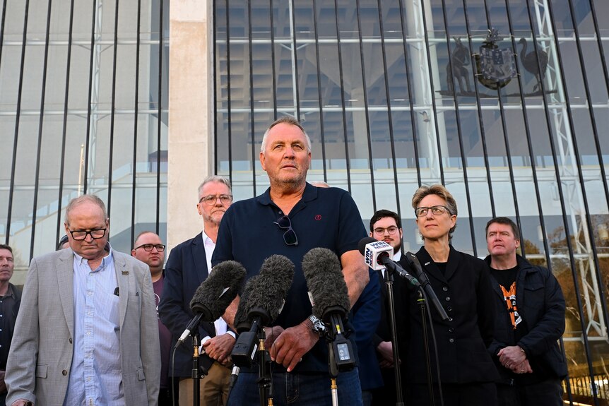 A man in a blue polo shirt stands in front of microphones with a group of people behind him at the High Court of Australia.