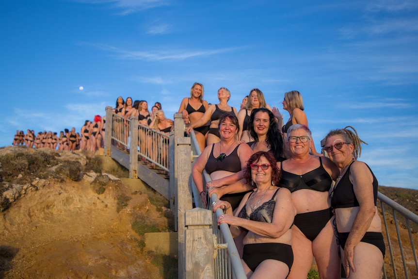 A large group of women, young and old, wearing black bras and undies, stand on a beach staircase and boardwalk