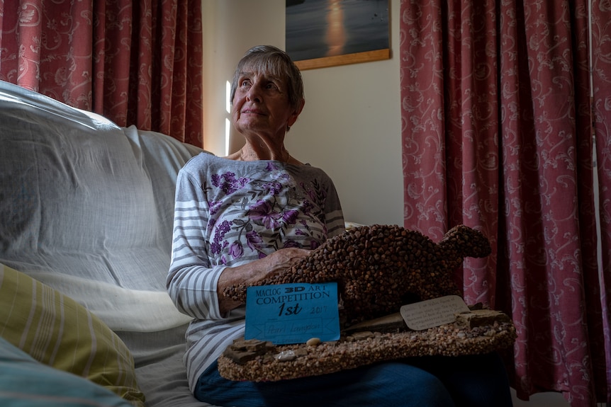 An elderly lady sits on a couch under a sunlit window holding a small artwork statue of a dinosaur made from stones.