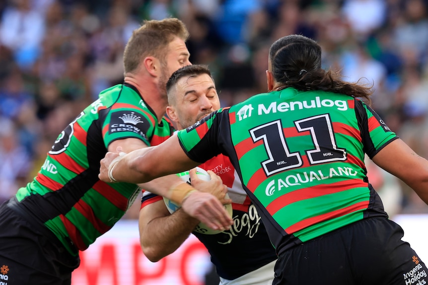 A Sydney Roosters NRL player holds the ball as he is tackled by two South Sydney opponents.