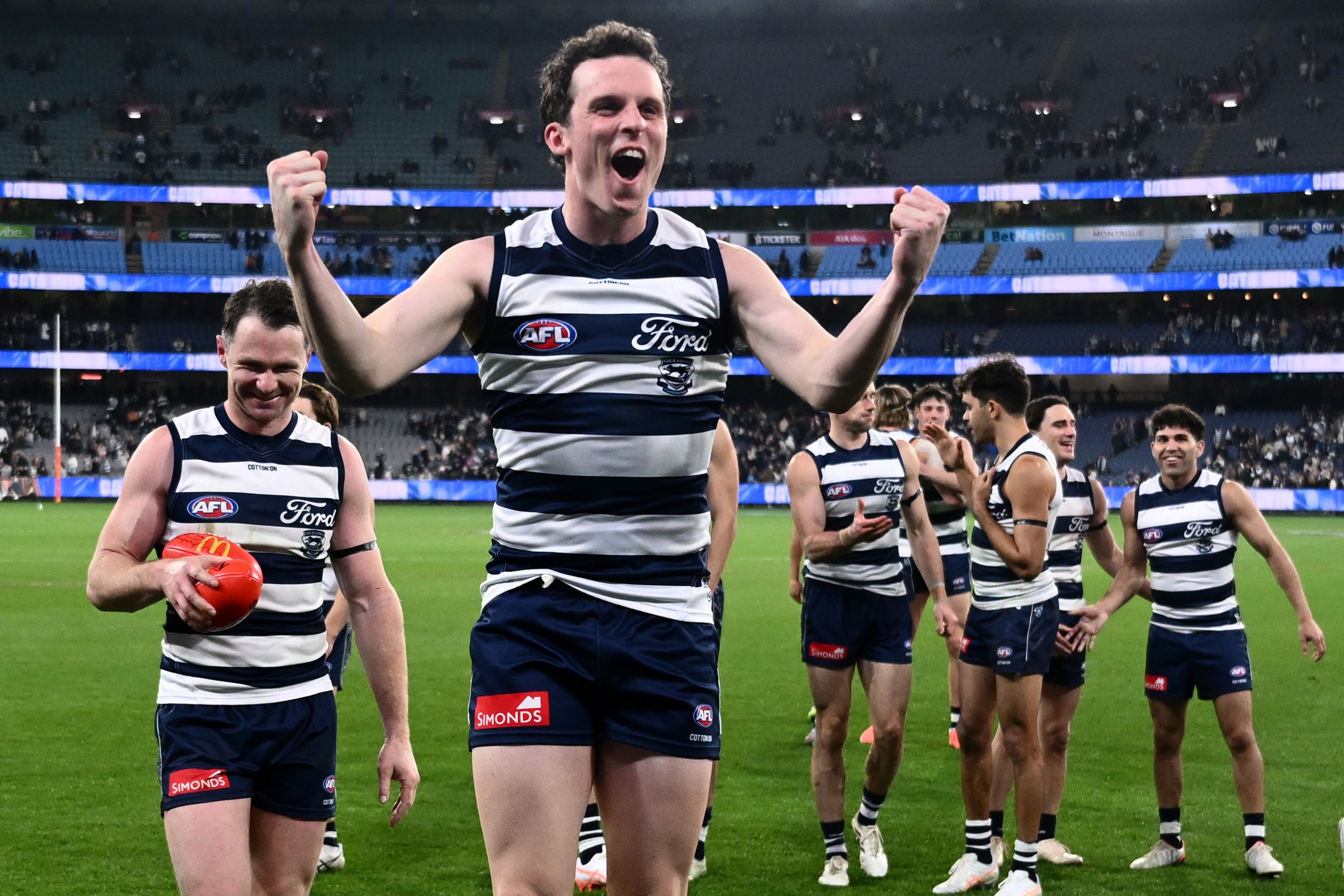 A Geelong AFL player grins and jumps off the ground in celebration with his fists pumped after a finals win.