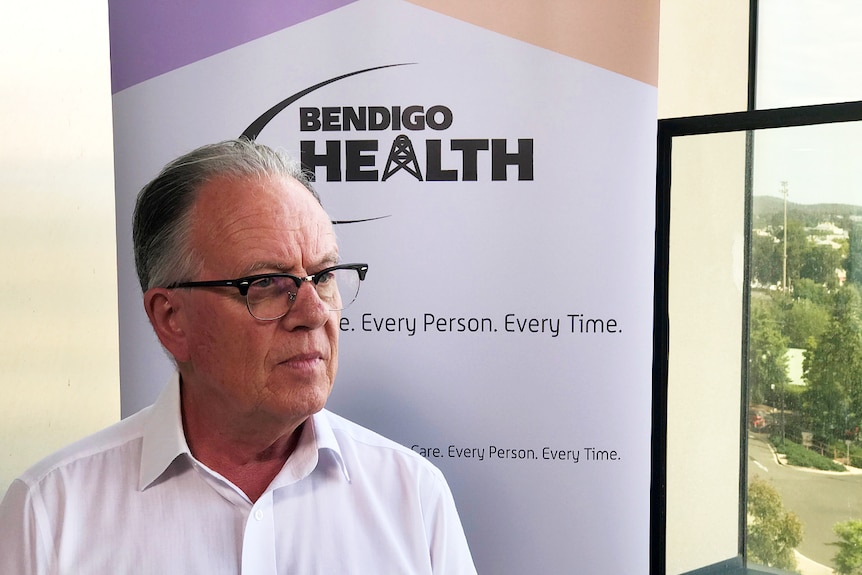 A grey man with glasses stands in front of a sign that says Bendigo Health