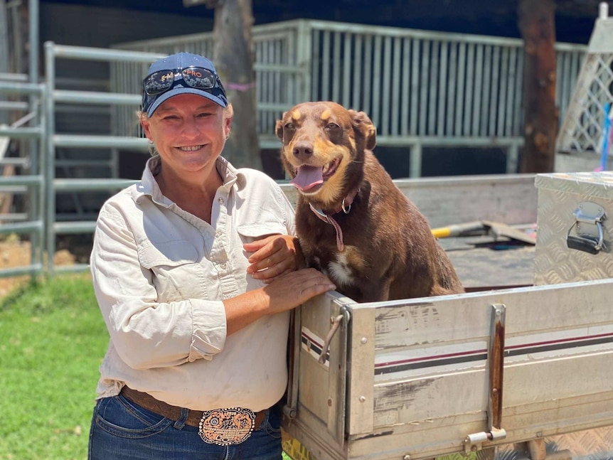 A woman stands smiling, next to her brown kelpie dog in the back of a ute.