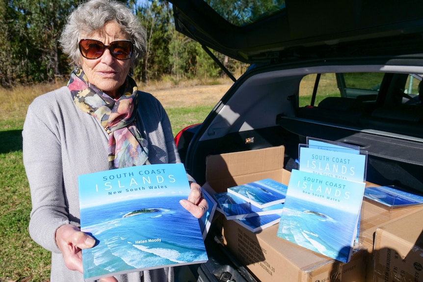 Elderly woman, short grey hair, grey top, multi-coloured scarf, dark glasses, holds book, South Coast Islands NSW, next to car.
