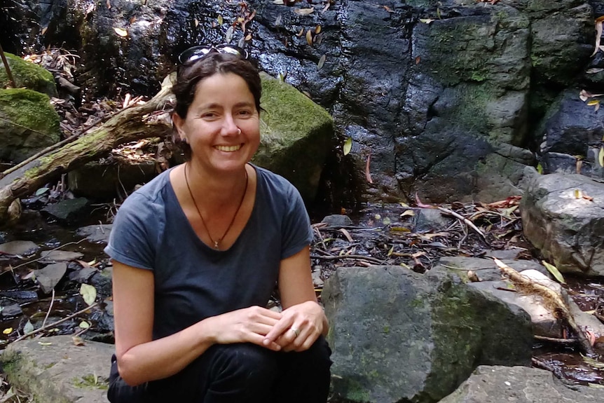 A lady smiling with a nose ring sits on moss-covered rocks with a stream trickling down the rockface behind her.