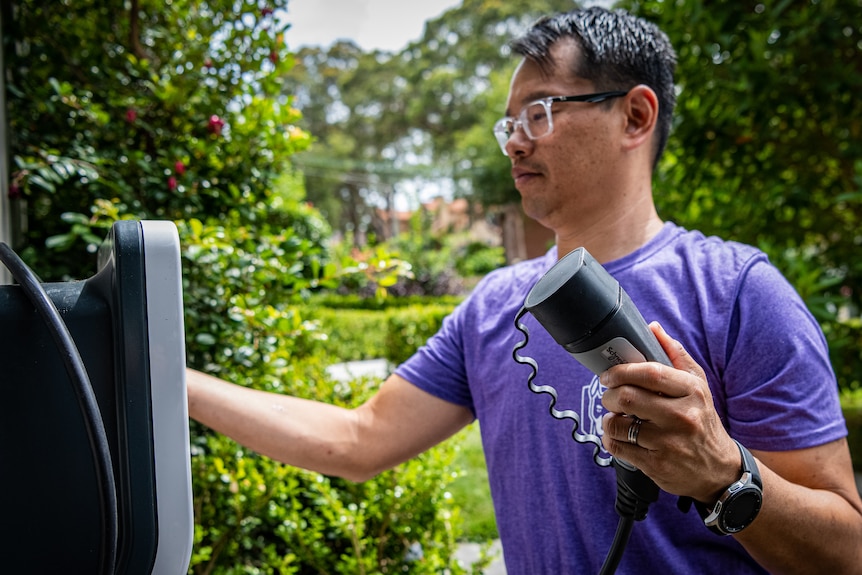 Marc Tan in a leafy front yard, holding an electric vehicle smart charger.