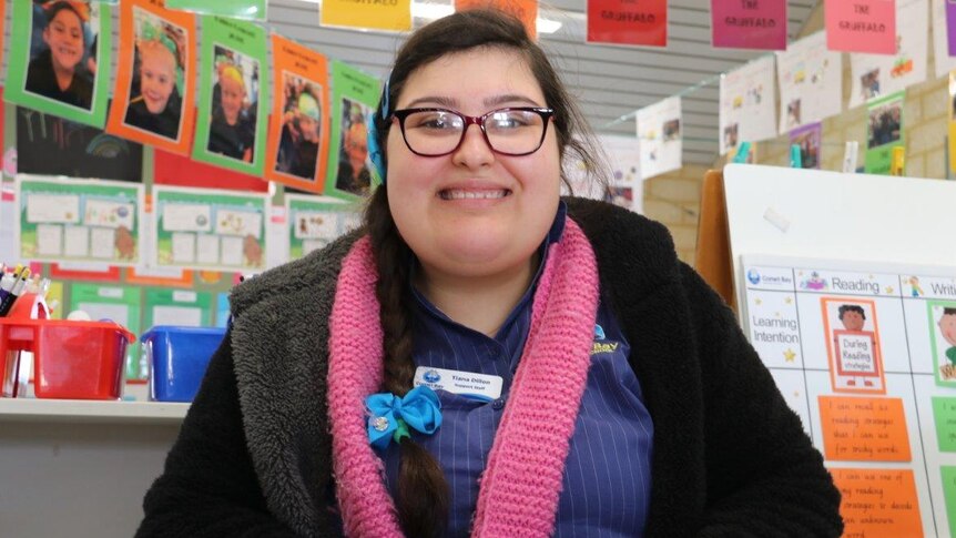 young teaching aide stands in primary school classroom smiling at camera