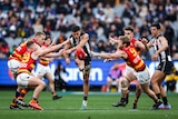 A Collingwood AFL player kicks the ball clear while four Crows players surrounding him try to smother the ball.