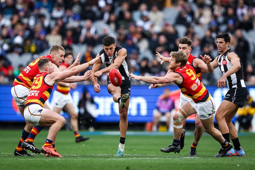 A Collingwood AFL player kicks the ball clear while four Crows players surrounding him try to smother the ball.