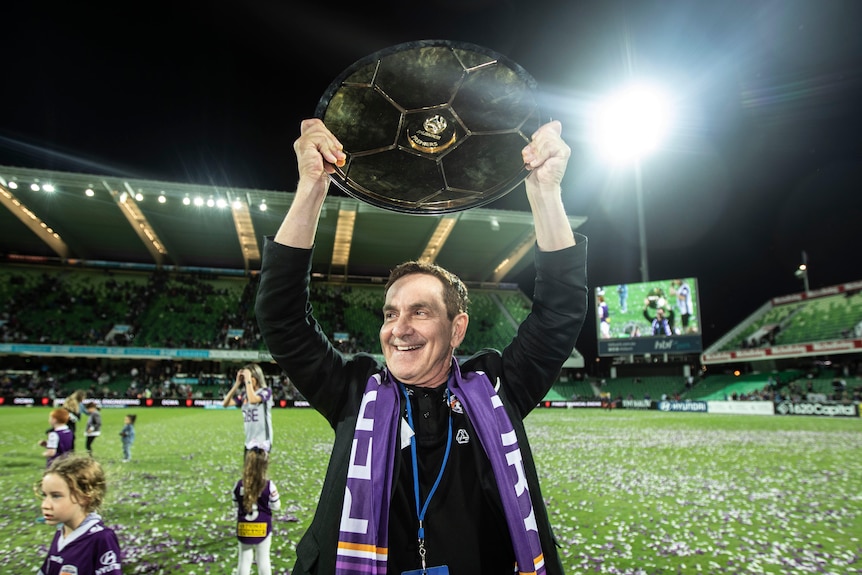 A man in a suit and a purple scarf celebrates on a pitch with a shield lifted over his head.