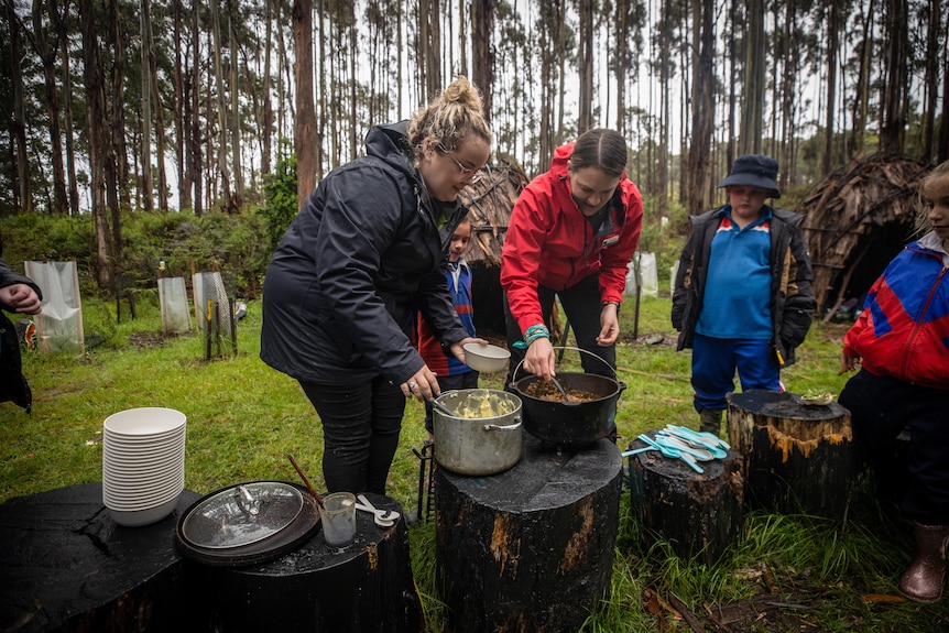 Two women in jackets ladle stew into bowels for a queue of children in a bush clearing.