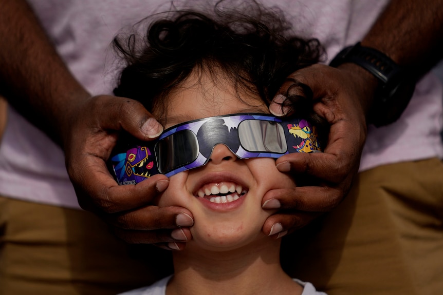 A child wearing eclipse glasses looks up at the sky