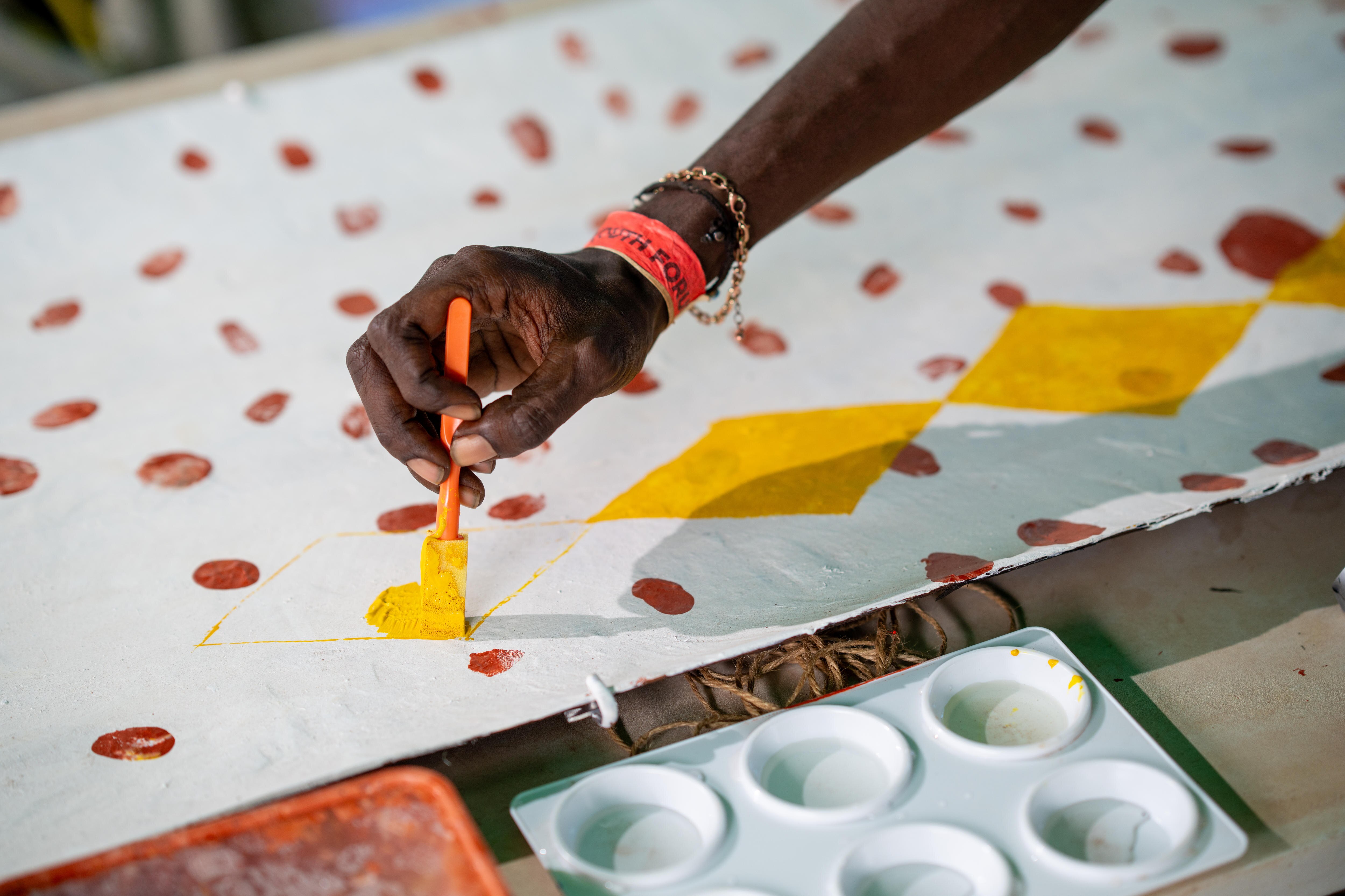Close up of a hand holding a paint brush, painting a yellow diamond on a white bark canvas. 