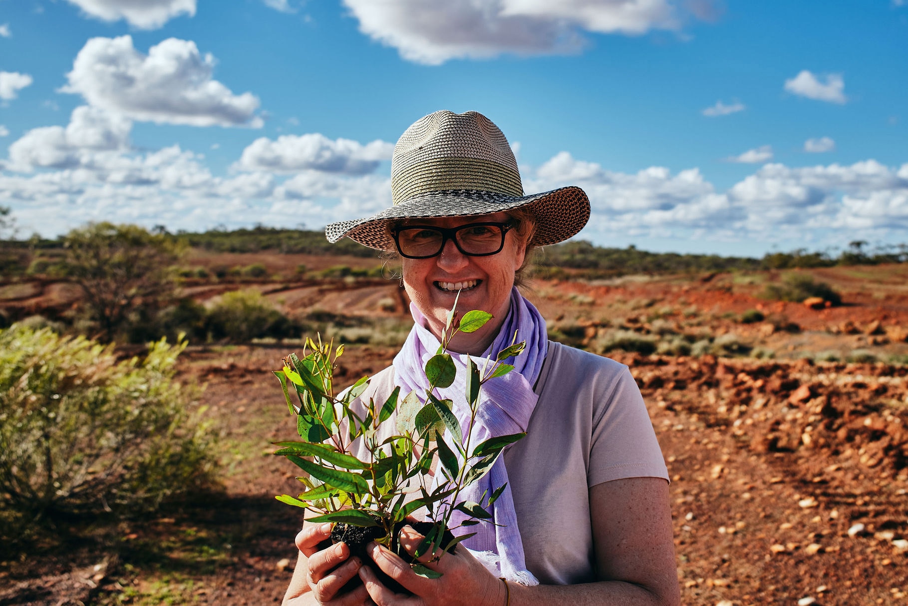 A smiling woman wearing a wide-brimmed hat and holding a plant stands outdoors in scrubland.