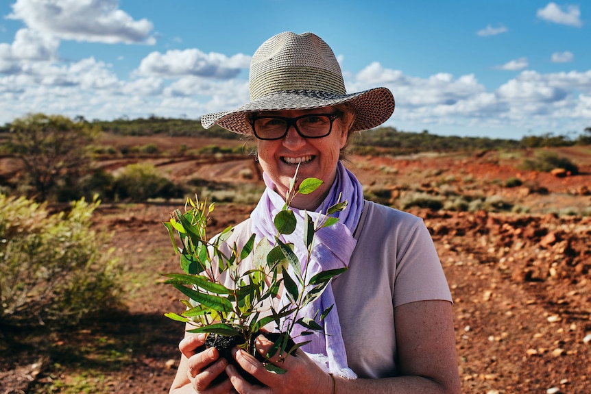 A smiling woman wearing a wide-brimmed hat and holding a plant stands outdoors in scrubland.