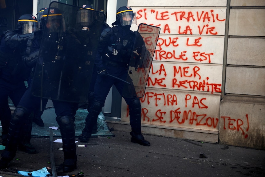 Black-clad riot police stand in front of a white stone building covered in red graffiti.