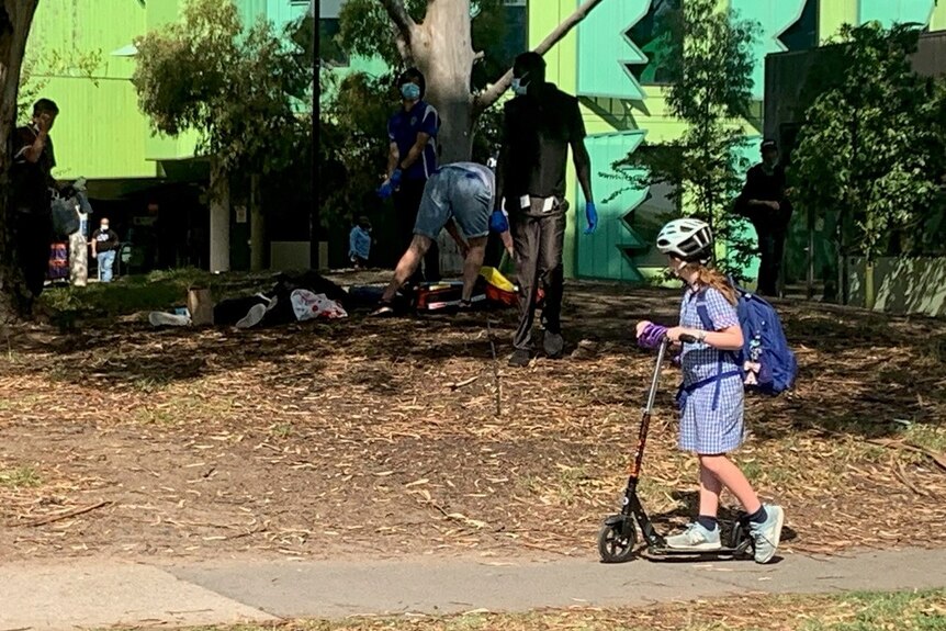 A child rides a scooter past a park where a drug user appears to be unconscious 