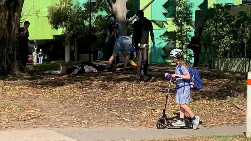 A child rides a scooter past a park where a drug user appears to be unconscious 