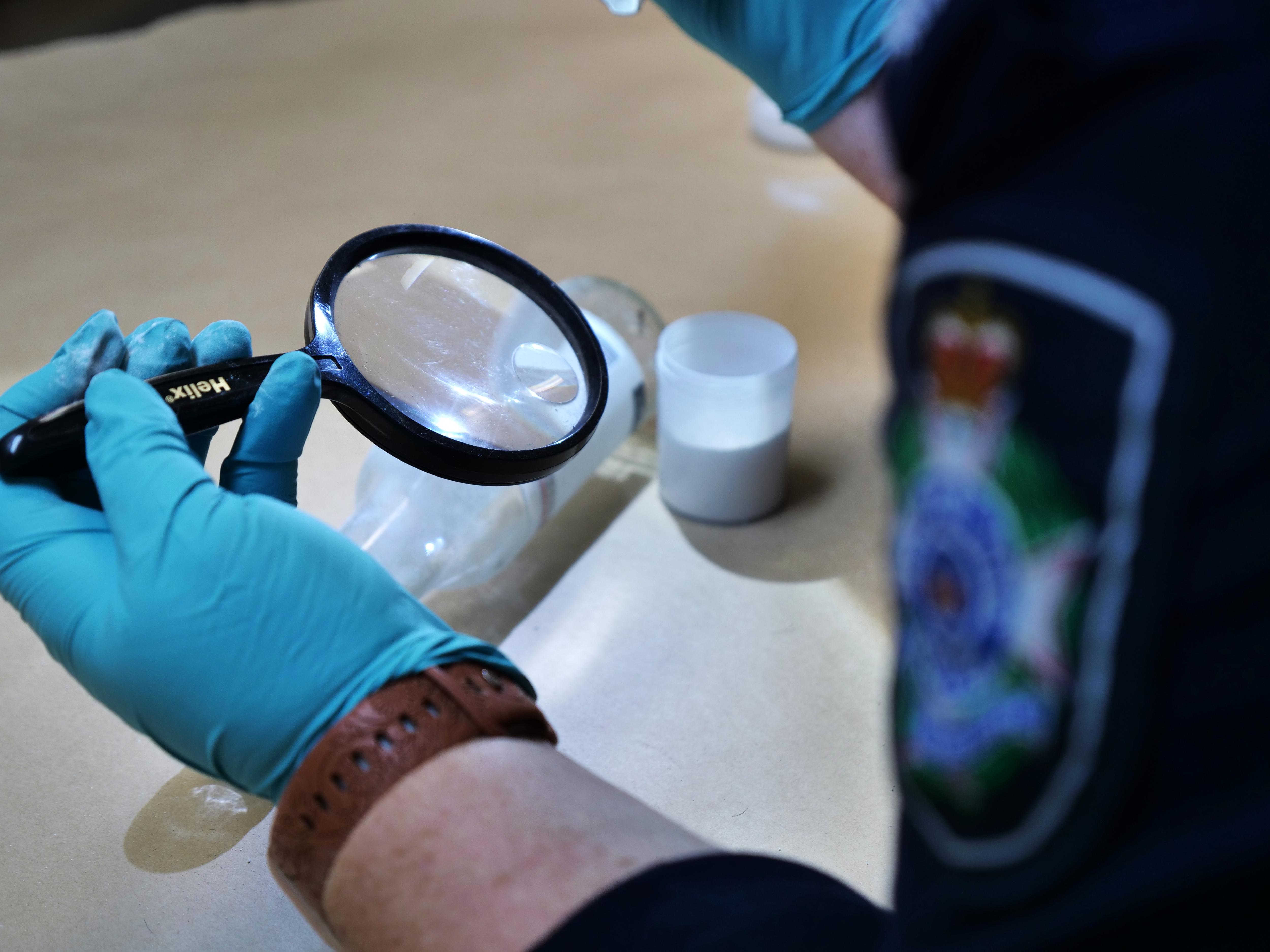 A hand with a blue glove holding a magnifying glass over a glass bottle. 