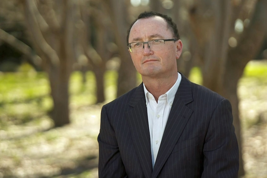 Man wearing a black suit jacket and white shirt, sitting in a park.