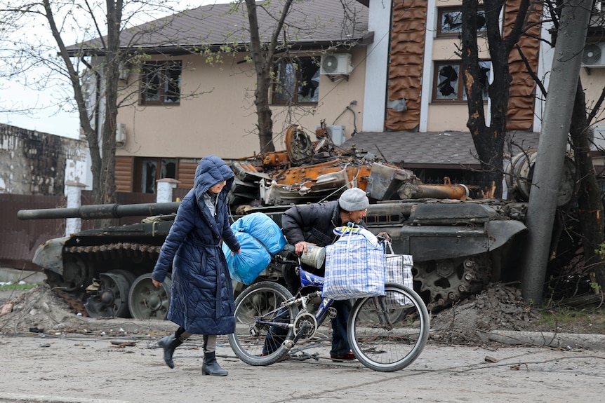 Two people walk with a large bag and a bicycle past a tank.