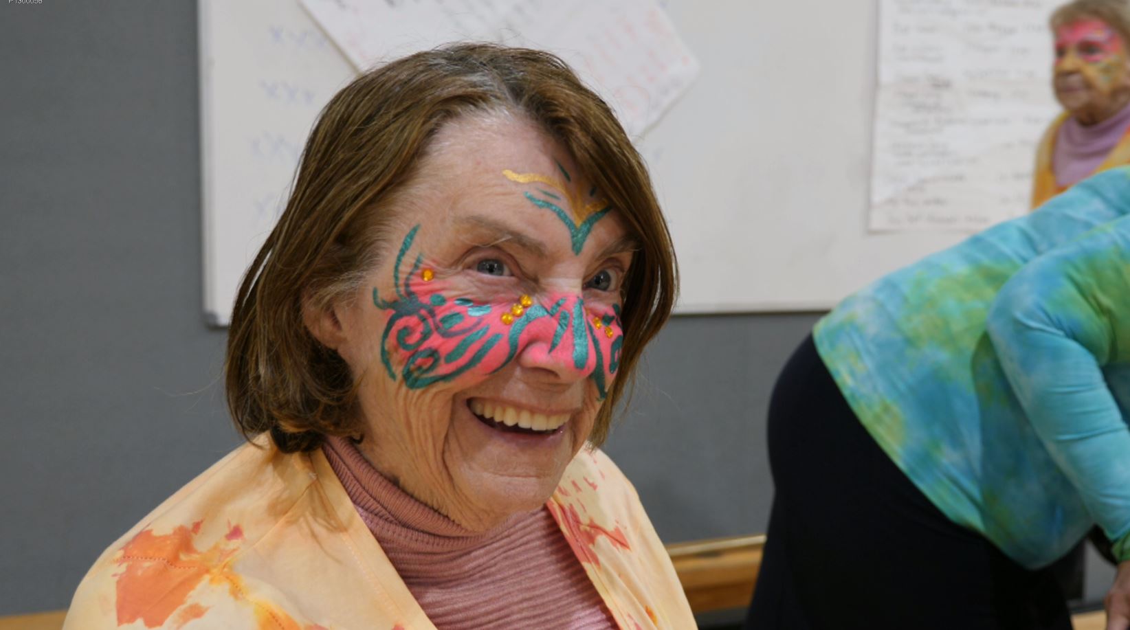 A woman in her 80s with tiedye yellow T-shirt, her face painted pink with green swirls across the cheeks and nose, smiling.
