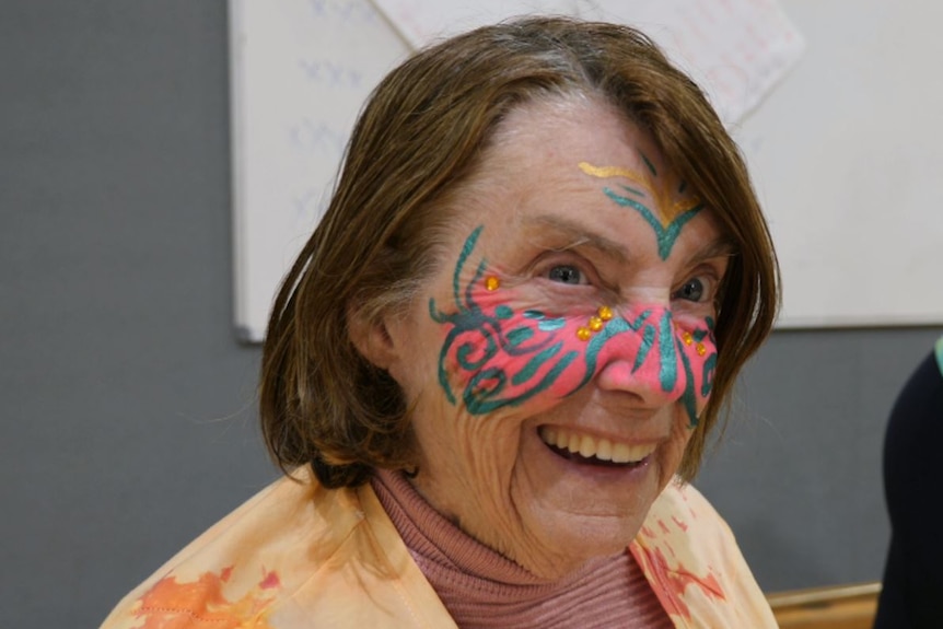 A woman in her 80s with tiedye yellow T-shirt, her face painted pink with green swirls across the cheeks and nose, smiling.
